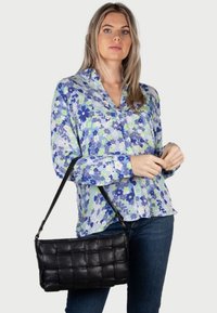 Zwarte geweven leren handtas met een rechthoekige vorm en schouderriem, gecombineerd met een bloemen blouse in blauw en groen en donkere jeans.