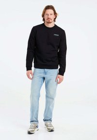 Felpa nera con collo a giro, polsini e orlo a costine; presenta un piccolo logo teal. Indossata con jeans blu chiaro e scarpe da ginnastica grigie.