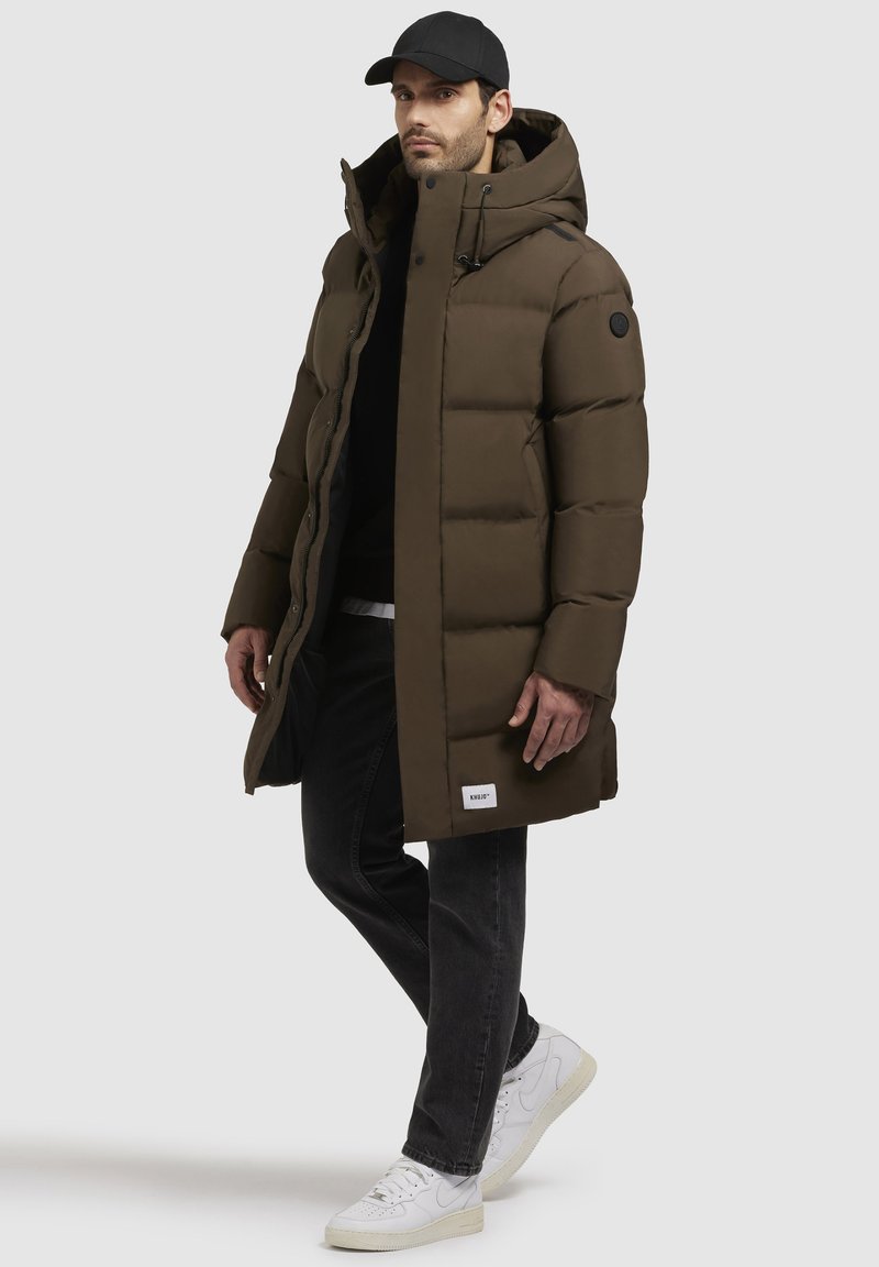 Doudoune marron avec capuche, présentant un design matelassé et des accents noirs. Portée sur un haut noir et associée à un jean foncé et des baskets blanches.
