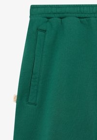 Pantalon de survêtement vert avec une taille élastique et une poche latérale verticale à gauche, doté d'une petite étiquette en tissu beige près de la poche.