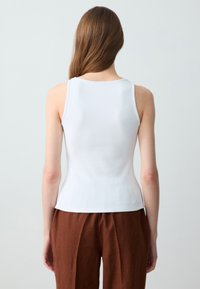 Weißes Rippentop mit rundem Halsausschnitt und breiten Schulterträgern, kombiniert mit lockeren braunen Leinenhosen. Glatte Textur.