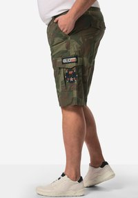 Shorts cargo en motif camouflage vert, avec de multiples poches, des écussons et une coupe ample. Associés à des baskets blanches.