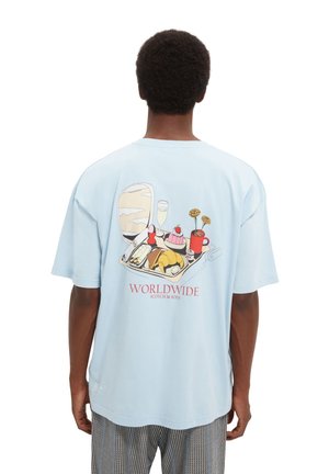 Persona che indossa una camicia azzurra chiara con una grafica colorata di un vassoio per pasti, fiori e il testo "WORLDWIDE Scotch & Soda" sul retro.
