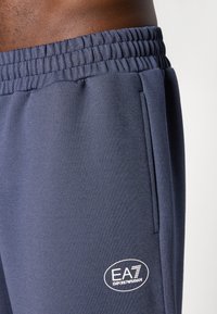 Ναυτικές αθλητικές φόρμες με υφή, ελαστική μέση, πλαϊνές τσέπες και το λευκό λογότυπο EA7 Emporio Armani στο κάτω μέρος του ποδιού.