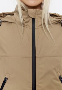 Beige gevoerde jas met een hoge kraag, zwarte rits en twee ritszakken. Heeft een gladde textuur en een afneembare capuchon.