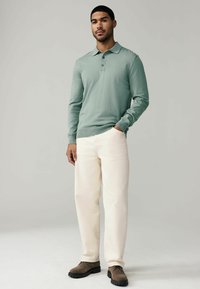 Hellgrünes Langarm-Polo-Shirt mit Kragen und drei Knöpfen; kombiniert mit cremefarbenen Hosen und braunen Schuhen. Glatte Textur.
