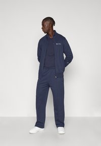 Μπλε ναυτικό φόρμα από ύφασμα sweatshirt, με μπουφάν με φερμουάρ και λογότυπο, μακριά μανίκια και ταιριαστό παντελόνι χαλαρής εφαρμογής. Άσπρα αθλητικά παπούτσια.