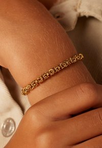 Gouden kettingarmband met ingewikkelde onderling verbonden ontwerpen, gedragen op een licht gebruinde arm, die een soepele metaaltextuur toont.