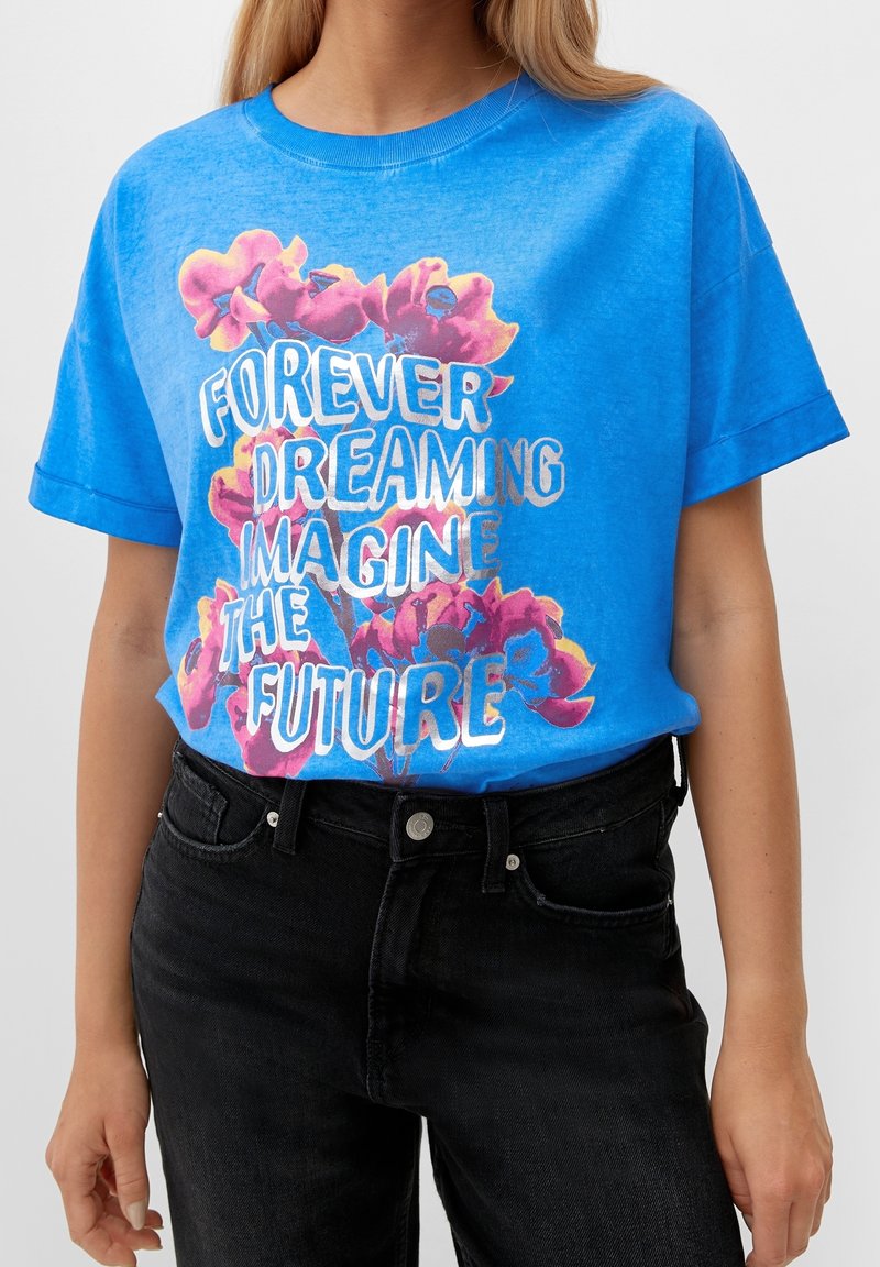 Femme portant un t-shirt bleu vif avec des fleurs roses et un texte blanc indiquant « Toujours rêver, imaginer le futur », associé à un jean noir.
