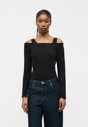 Žena s copánky, která nosí černý top s dlouhým rukávem a odhalenými rameny a tmavě modré džíny, stojící před jednoduchým bílým pozadím.