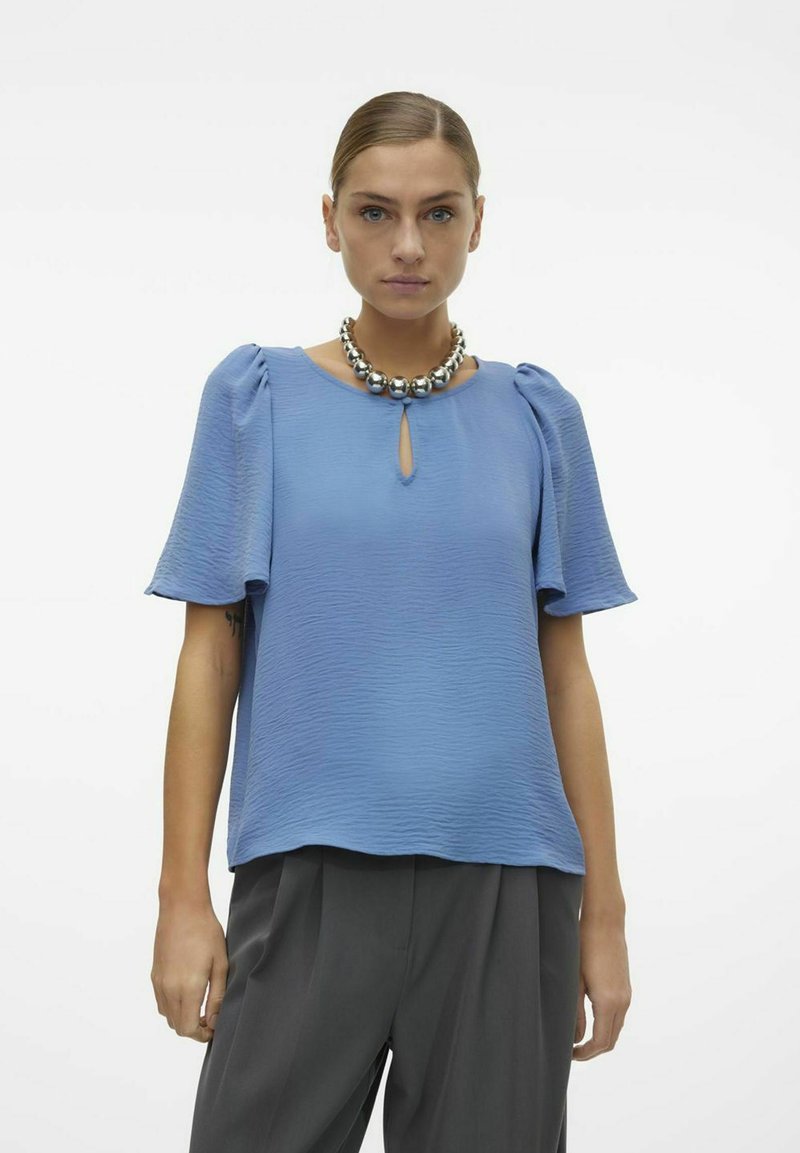 Blusa azzurra a maniche corte con tessuto testurizzato, scollatura a chiave e maniche a sbuffo, abbinata a una collana argentata voluminosa.