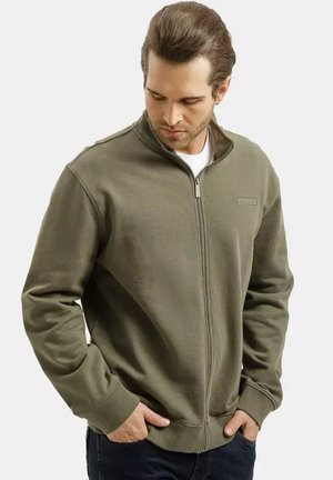 Mężczyzna w oliwkowej, w pełni zapinanej kurtce z wysokim kołnierzem, z rękami w kieszeniach, patrzący w dół, stojący na jednolitym tle.