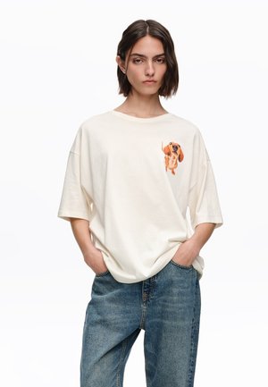 Joven con cabello oscuro hasta los hombros, lleva una camiseta blanca holgada con un gráfico de un perro salchicha y jeans azules, manos en los bolsillos, expresión neutral.