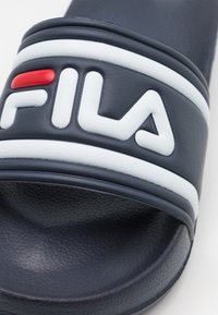 Marinblå slidesandaler med vita och röda accenter. Har en texturerad yta och en framträdande upphöjd "FILA"-logotyp på remmen.