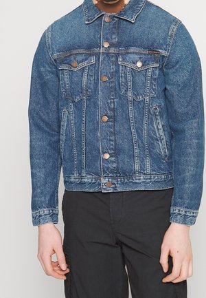 Mężczyzna w niebieskiej kurtce dżinsowej z zapięciami na guziki i czarnych spodniach, stojący na jasnym, jednolitym tle.