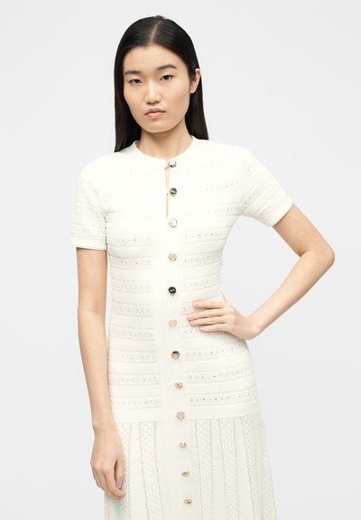 Γυναίκα με μακριά σκούρα μαλλιά που φοράει λευκό, κοντομάνικο, πλεκτό φόρεμα με κουμπιά και υφασμένους οριζόντιους και κάθετους μοτίβους.