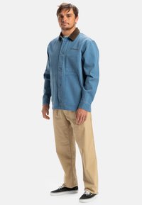 Blue denim jacket with brown collar, button front, and two chest pockets, paired with beige pants and black sneakers. Casual layered look.