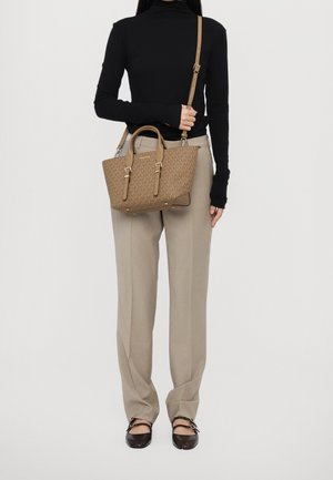 Personne portant un haut à manches longues noir et un pantalon beige tenant un sac à main marron à motifs avec deux sangles et des boucles dorées.