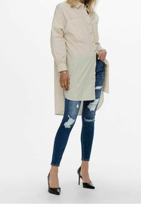 Knopfhemd mit langen Ärmeln in hellem Beige und längerem Rückenteil, kombiniert mit abgewetzten blauen Jeans und schwarzen High Heels.