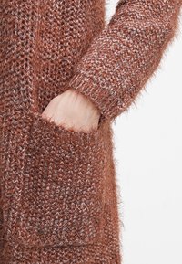 Brun stickad kofta med en mjuk, luddig yta, långa ärmar och en synlig ficka. Tyget har en blandning av ljusa och mörka trådar.