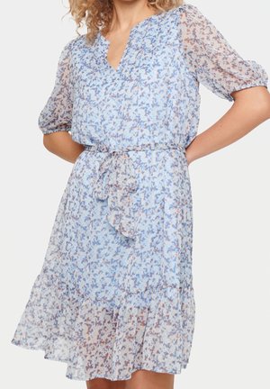 Jasnoniebieska sukienka w kwiaty z przezroczystymi bufkami na rękawach, paskiem wiązanym w talii i falbaną u dołu, noszona przez osobę z kręconymi włosami.