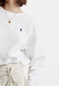 Person klädd i en vit sweatshirt med en liten marinblå polo-spelare logo och beiga högmidjade byxor med en knuten bälte.