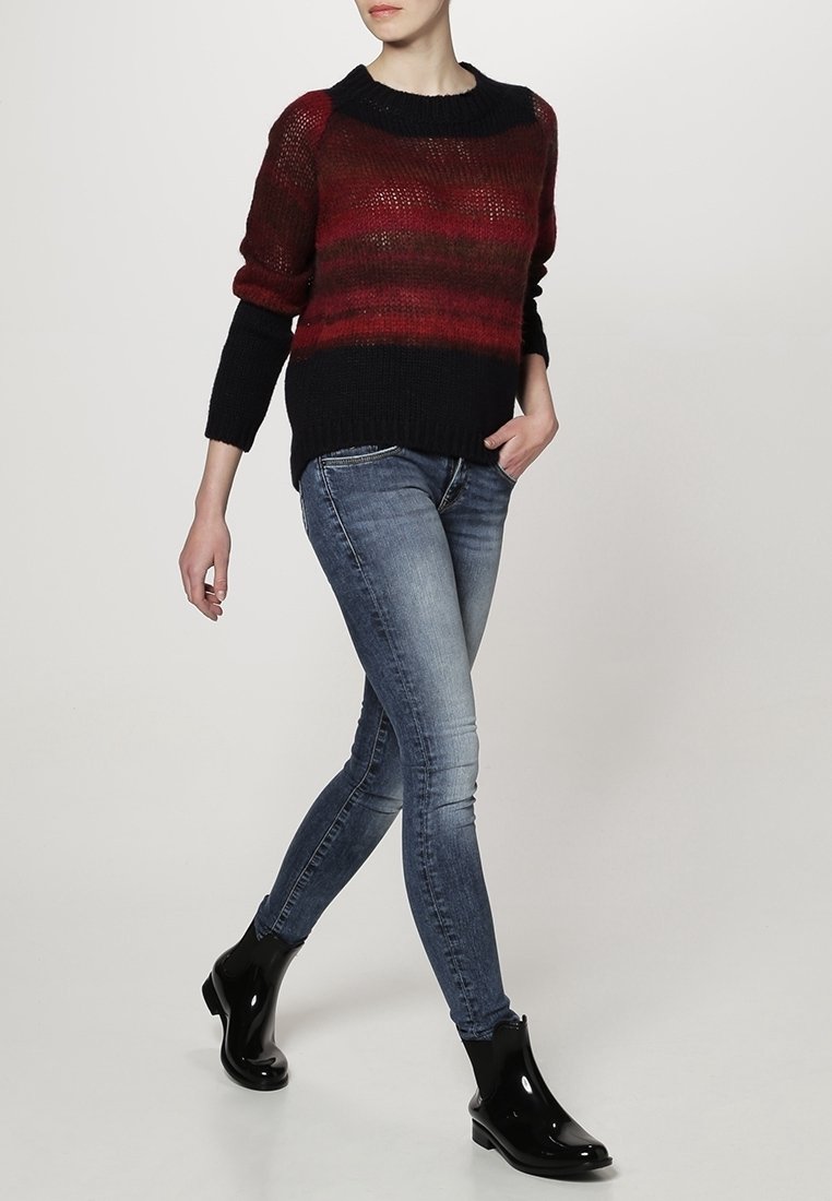Personne portant un pull dégradé rouge et noir, un jean skinny bleu délavé et des bottines noires brillantes, debout avec une main dans la poche.