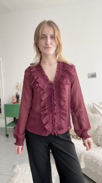 Burgundy paita, jossa on rypytetty etuosa, V-pääntie ja pitkät hihat. Läpinäkyvä kangas, teksturoitu hihansuu, yhdistetty mustiin housuihin.