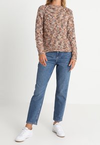 Pletený svetr v teplých odstínech s texturovaným vzorem a pružným lemem, kombinovaný s džíny rovného střihu modré barvy a bílými teniskami.