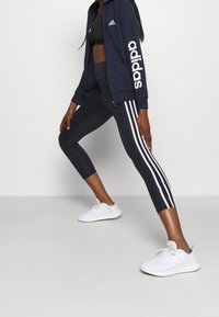 Marinblå zip-up huvtröja med vit "adidas" text, kombinerad med svarta leggings med vita sidränder, och vita träningsskor.