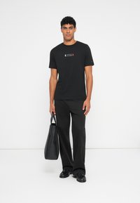 Μαύρη μπλούζα με εκτυπωμένο λογότυπο σε κόκκινο, άσπρο και μπλε χρώμα στο στήθος. Το μοντέλο φοράει μαύρο παντελόνι και κρατάει μια μαύρη τσάντα tote. Υφασμάτινη υφή.