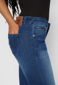 Pantalones vaqueros de mezclilla azul oscuro con una textura suave, que presentan una etiqueta de cuero con la inscripción "TOMMY JEANS" y un diseño de cinco bolsillos con costuras en contraste.