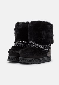 Botines tobilleros de piel sintética negra con base gruesa, adornados con cadenas metálicas y lados con patrones. Diseño texturizado y acolchado.