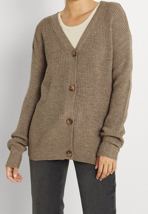 Cardigan tricoté marron à quatre boutons porté sur un haut beige, associé à un pantalon gris foncé, visible de la poitrine à mi-cuisse.