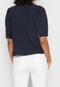 Blouse bleu marine à manches courtes avec épaules bouffantes, présentant des accents de boutons et une coupe décontractée, associée à un pantalon en denim blanc.