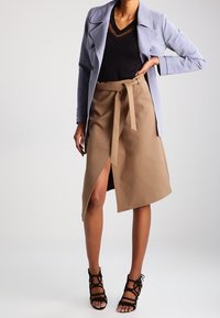 Kvinna som bär en ljusblå jacka, svart V-ringad topp, beige omlottkjol med slits och svarta remsandaler med hög klack, ståendes med en hand på höften.