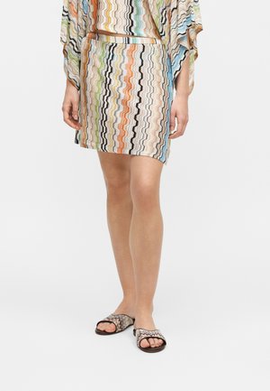 Nő visel egy több színből álló cikk-cakk mintás szoknyát és hozzáillő felsőt, fekete slide szandálokkal, amelyek hasonló cikk-cakk mintával rendelkeznek.