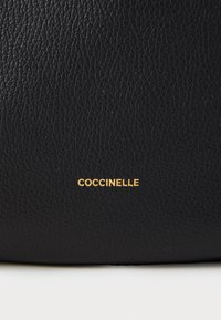 Fekete, texturált bőr táska, amelyen az alján egy arany "Coccinelle" logó található, hangsúlyozva a sima, tartós anyagot és a tömör márkázást.