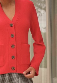 Red cardigan made of knit fabric, featuring a v-neck, five brown buttons, and two front pockets. Long sleeves with a fitted silhouette.