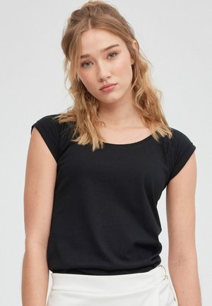 Joven mujer con cabello rubio ondulado hasta los hombros, vestida con una camiseta negra de manga corta y falda blanca, expresión neutral, fondo gris claro.