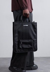 Black nylon tote bag with top handles, featuring a front zip pocket and a minimalist logo, rectangular shape, and smooth texture.
