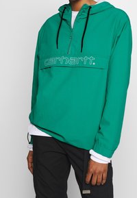 Blågrøn pullover-jakke med en halv lynlås og et logo-panel foran. Letvægtsstof, justerbar hætte og sorte snore.