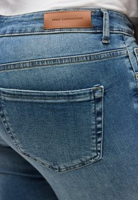 Κοντινή λήψη πίσω τσέπης από μπλε τζιν με δερμάτινη ετικέτα μάρκας "ONLY CARMAKOMA" στη μέση, φορεμένο με λευκή μπλούζα.