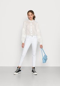 Camisa branca transparente com mangas bufantes, botões pretos, jeans skinny brancos, ténis altos pretos e uma mala estruturada azul clara.