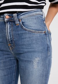 Džínové kalhoty světle modré barvy s kamenovaným povrchem. Obsahují patche s opotřebením a kontrastní oranžové švy. Klasický design s pěti kapsami.