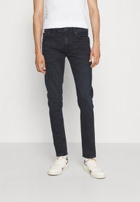 Dark blue denim jeans with a slim fit, five-pocket design, visible stitching, and a smooth texture, paired with white sneakers.