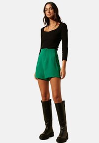 Haut noir à manches longues côtelé associé à un short vert taille haute ; le mannequin porte des bottes noires brillantes à hauteur de genou avec des semelles épaisses.