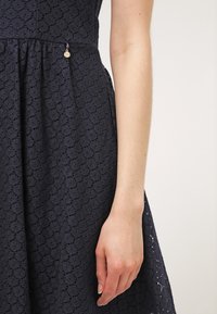 Navyblaues Spitzenkleid mit strukturiertem Muster, schwingendem Rock und seitlichem Akzent mit einem kleinen metallic Anhänger. Eine Hand ist teilweise sichtbar.