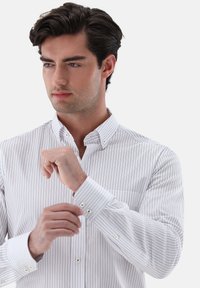 Weißes Hemd mit feinen, vertikalen Marineblauen Streifen. Verfügt über einen klassischen Kragen, verstellbare Manschetten mit zwei Knöpfen und einer Brusttasche.