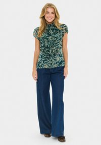 Lächelnde Frau mit langen blonden Haaren, die eine durchsichtige, kurzärmelige grüne Bluse mit Blumenmuster, weit geschnittene blaue Jeans und braune Schuhe trägt.